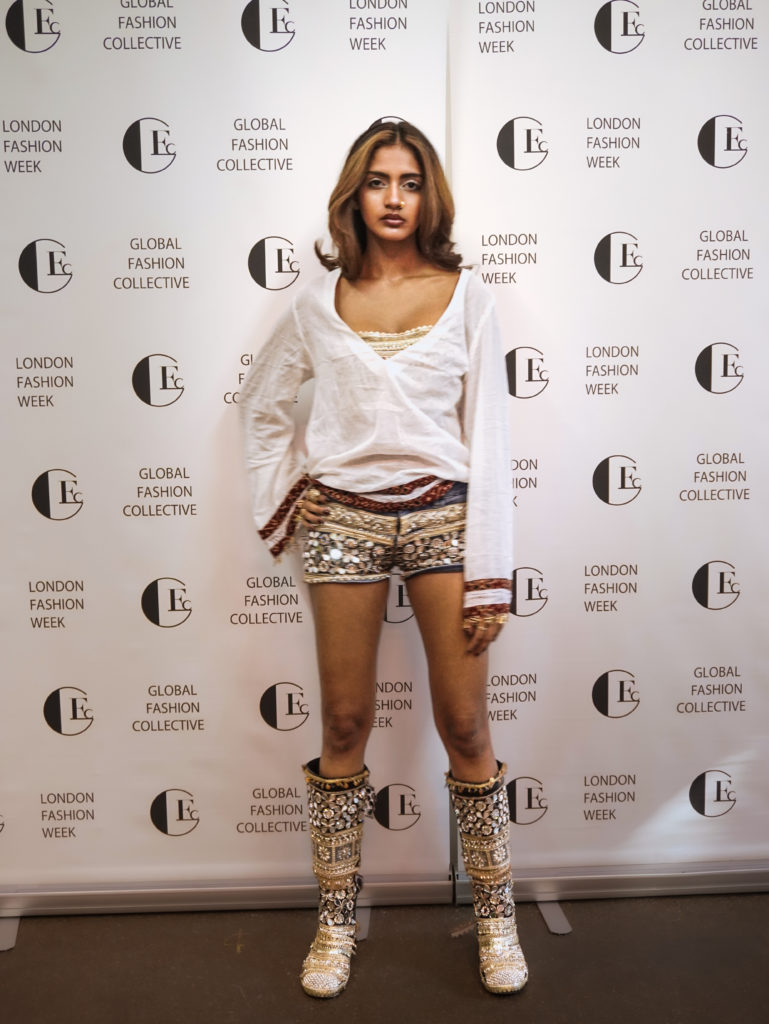 Model posing in front of a London Fashion Week backdrop with Global Fashion Collective logos, wearing a white wrap top, patterned shorts, and embellished knee-high boots.