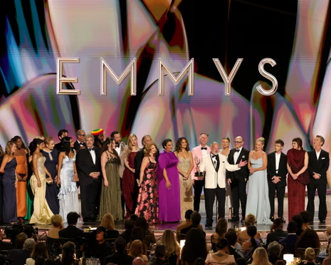 Group of Emmy Award nominees and presenters on stage in formal attire under a large'EMMYS' backdrop, with an audience visible in the foreground.