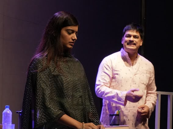 Sakshi Singh - Social Nation Two performers on a dimly lit stage: a woman in a dark patterned outfit looks down, while a man in a light pink shirt gestures beside her.